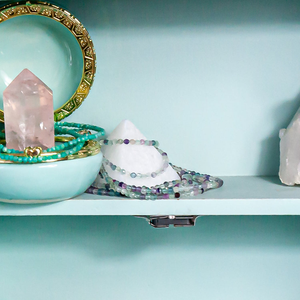on the left, 2mm amazonite faceted crystal necklace around rose quartz point inside jewelry box. in the middle 2mm fluorite faceted crystal necklace around selenite pyramid - all inside jewelry cabinet.
