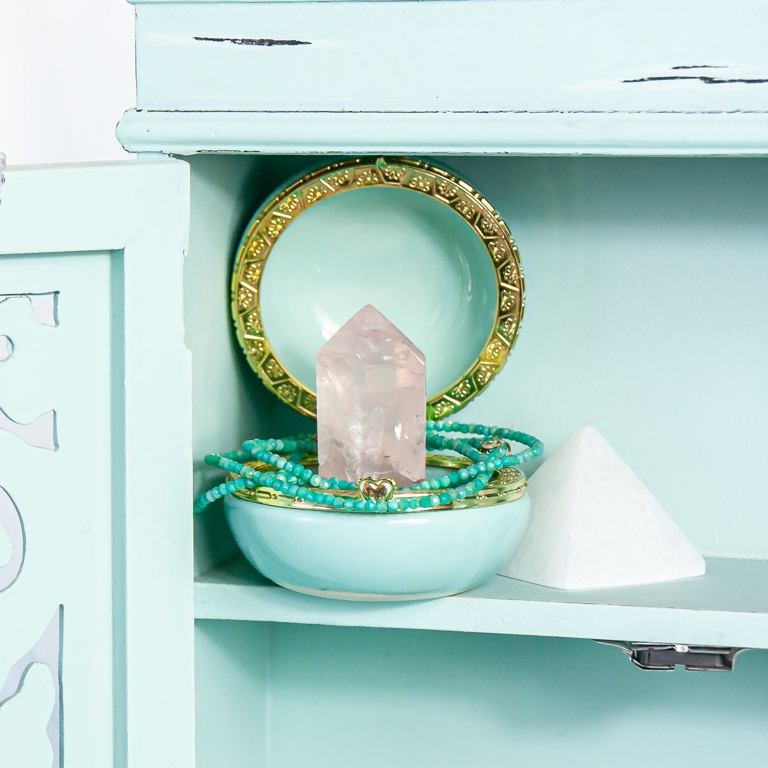 2mm amazonite faceted crystal necklace set around rose quartz point inside jewelry box. to the right of it sits a selenite crystal pyramid. all inside cabinet. close-up.