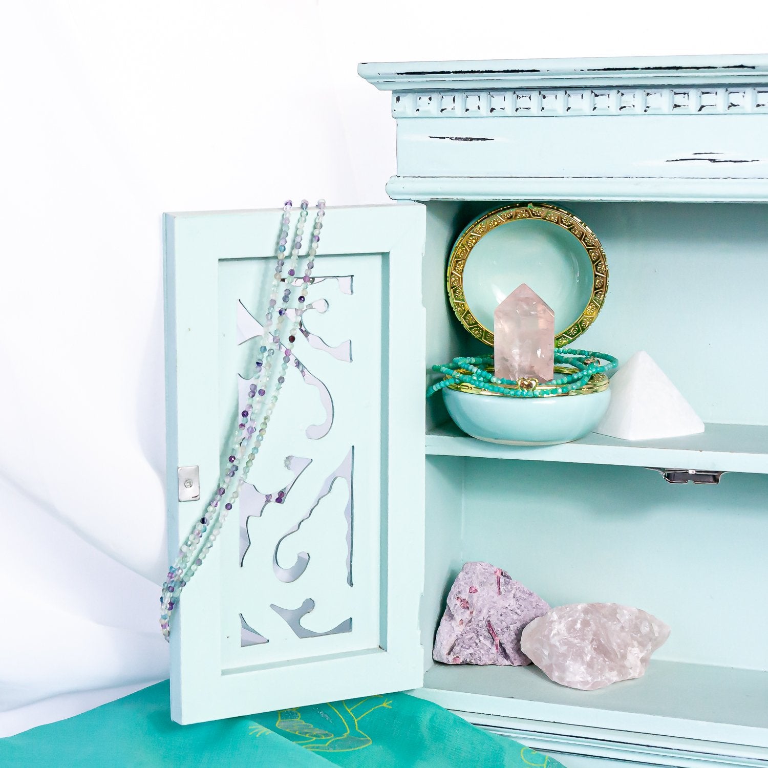 on the left, 1mm fluorite faceted crystal  bead necklace on cabinet door; on the right, 2mm amazonite faceted crystal bead necklace  set around rose quartz point in jewelry box. also in cabinet for aesthetics are a selenite crystal pyramid, raw pink tourmaline rock and raw rose quartz crystal rock.